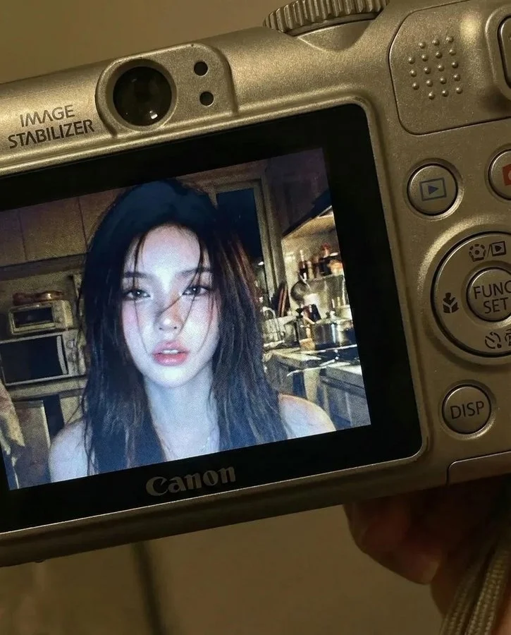 AI image: Close-up shot of young woman displayed on compact Canon digital camera screen. Camera body surrounds image with visible buttons, dials, textured surface. Photo shows woman indoors at night illuminated by bright built-in flash creating sharp highlights on face and hair. Long dark hair falling across face in loose strands. Candid, raw, nostalgic early-2000s digital camera snapshots. Slightly muted colors with cool undertones, strong flash contrast, natural grain.
